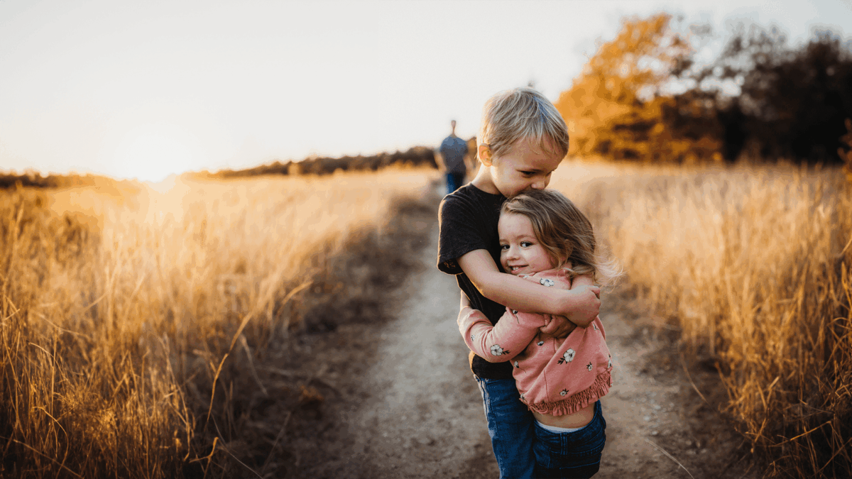 Symbolbild: Ein blonder Bub umarmt ein blondes Mädchen. sie stehen gemeinsam auf einen Feldweg und im Hintergrund ist ein Sonnenuntergang zu sehen.