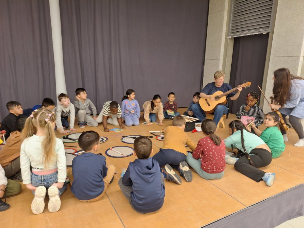 Eine Gruppe von Kindern sitzt mit zwei Musikerinnen auf der Bühne in einem Kreis.