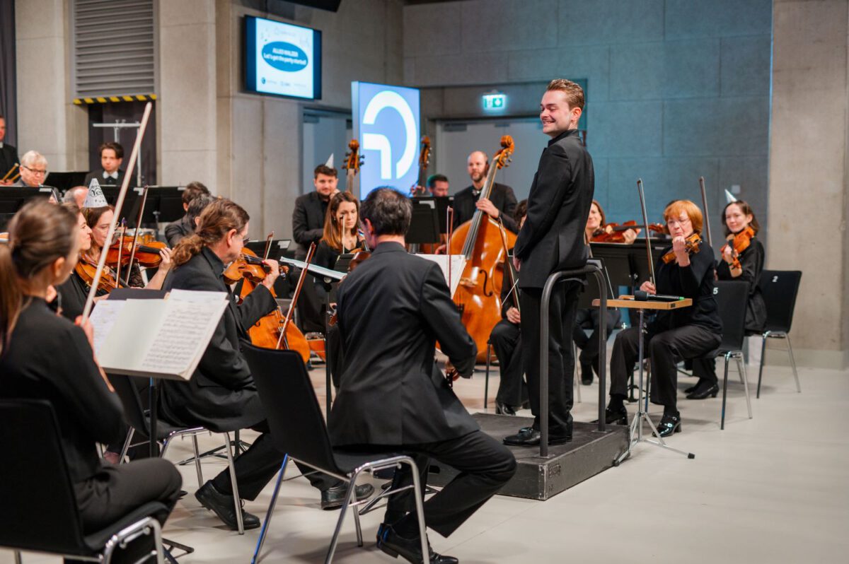 Ein Dirigent steht auf einem Dirigentenpult und vor ihm sitzt ein Orchester.