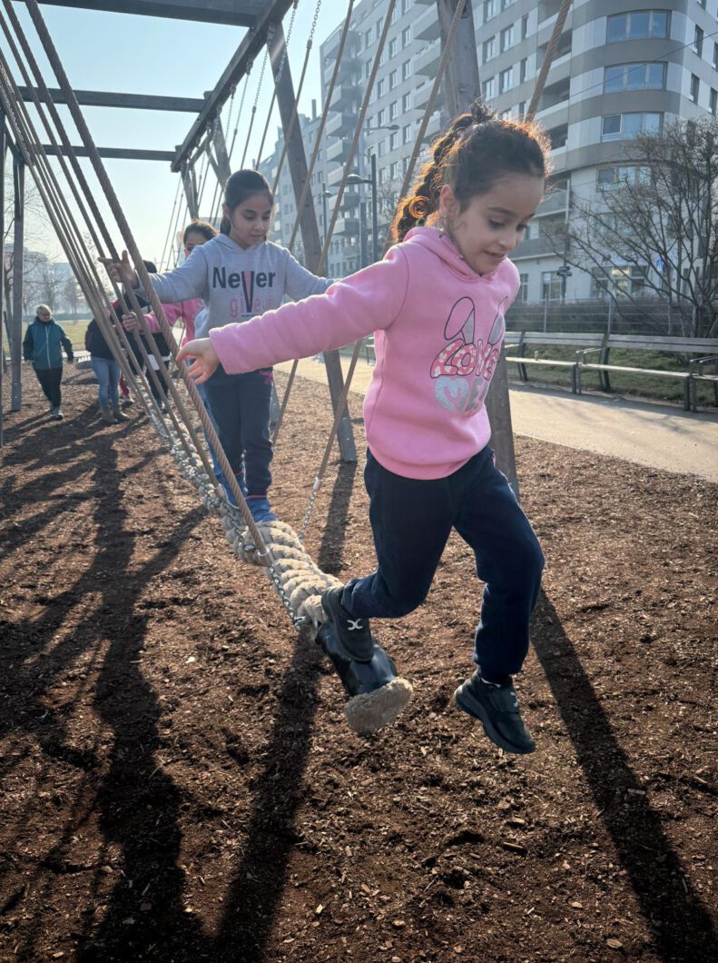 Zwei Mädchen balancieren auf einem Seil auf einem Kinderspielplatz. Ein Mädchen springt von
dem Seil auf den Boden.