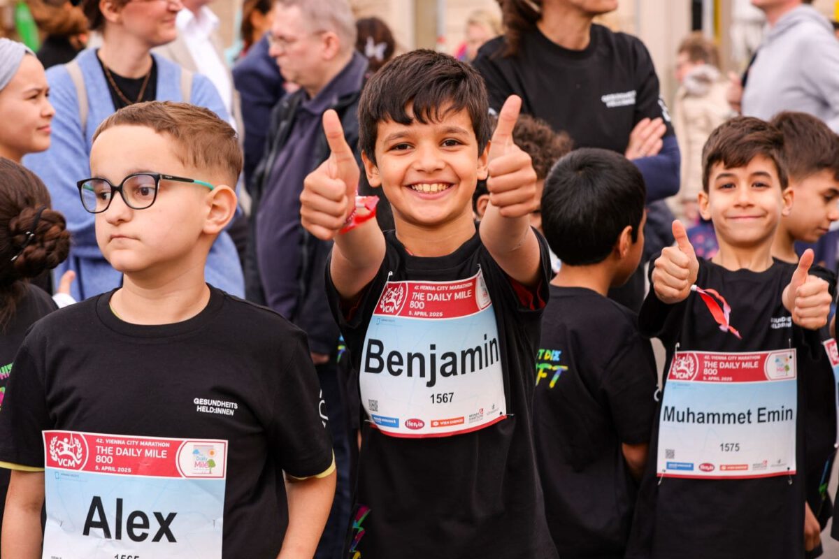 Drei lächelnde Kinder mit Startnummern stehen beim Vienna City Marathon in der Menge. Zwei Jungen strecken beide Daumen hoch.