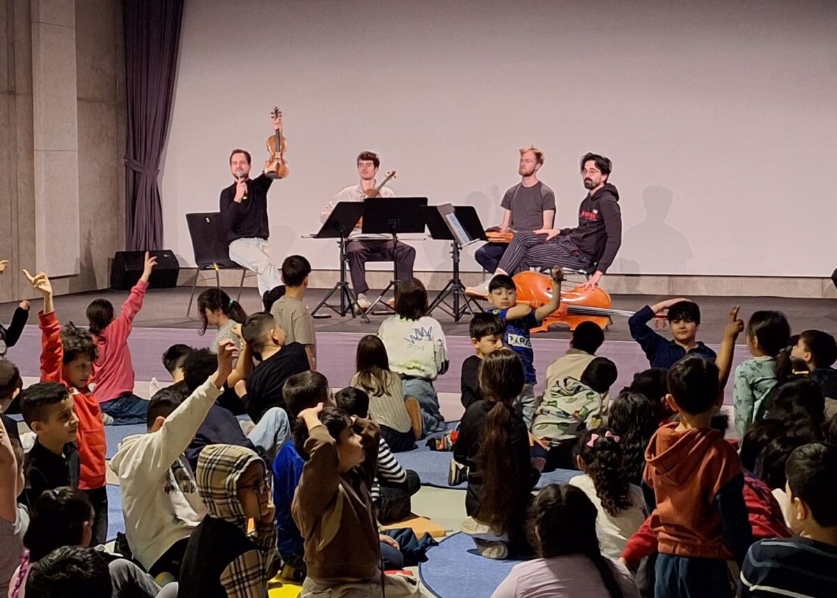 Ein Streichquartett tritt auf der Bühne im CAPE 10 vor einigen sitzenden Kindern auf. Einer der Musiker hält eine Geige in die Höhe.