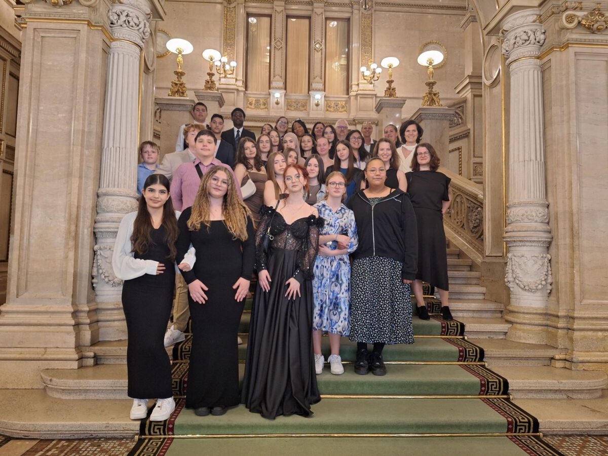 Eine Gruppe junger Menschen in festlicher Kleidung posiert auf einer prunkvollen Treppe in der Wiener Staatsoper.