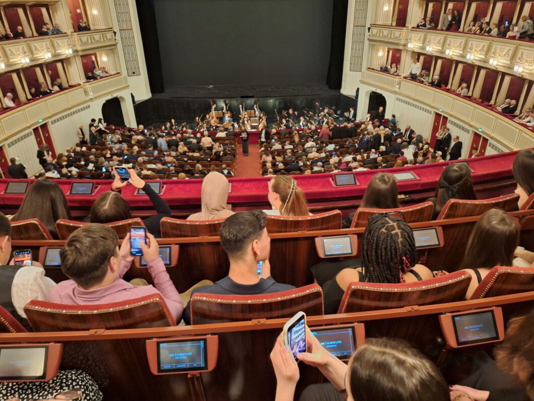 Eine Gruppe von Schüler:innen (von hinten gesehen) 
sitzt in der Mitteloge der Wiener Staatsoper mit Sicht auf das Orchester.