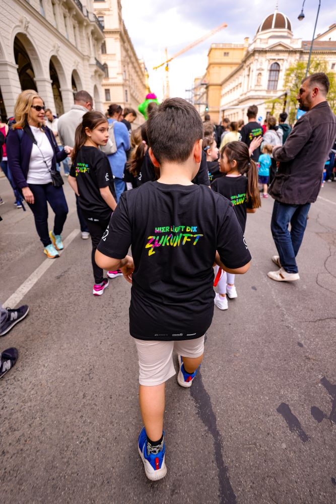 Eine Gruppe von Kindern (von hinten gesehen) läuft in schwarzen Shirts mit der bunten Aufschrift „Hier läuft die Zukunft“ beim Vienna City Marathon mit.