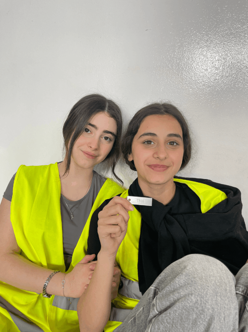 Zwei Mädchen sitzen an einer Wand. Beide lächeln und 
eine hält ein Schild mit der Aufschrift „TINA“ in der Hand.