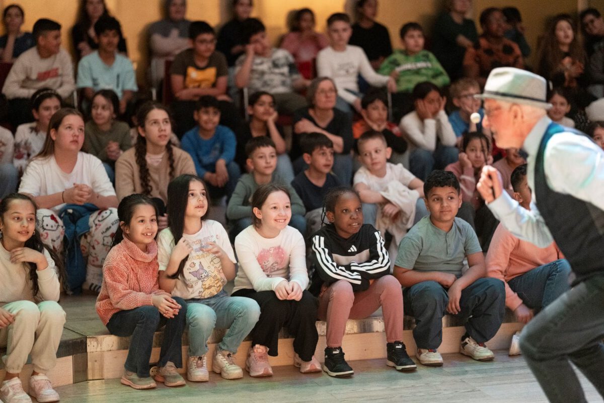 Musikvermittler Albert Landertinger performt auf der Bühne und sieht in Richtung des lachenden Kinder-Publikums.