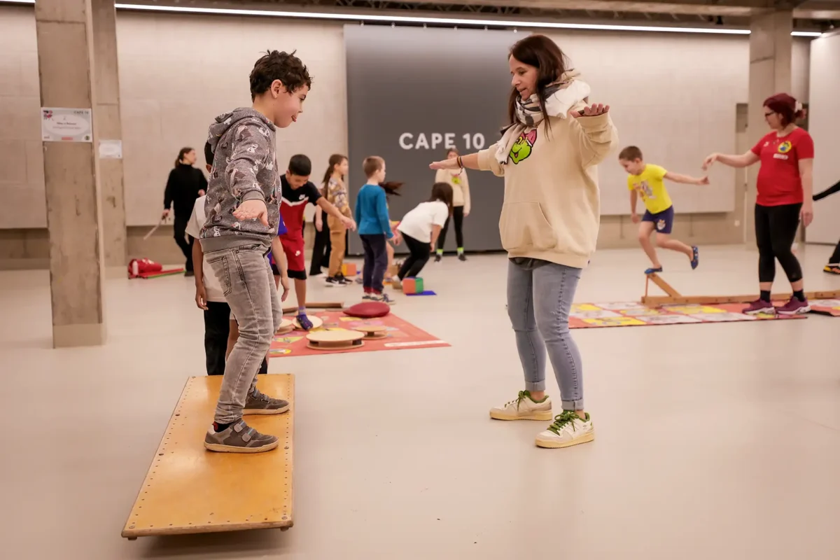 Ein Bub steht mit auf einem Balance-Board, gegenüber von ihm steht eine Betreuerin und zeigt ihm wie die Balance hält.