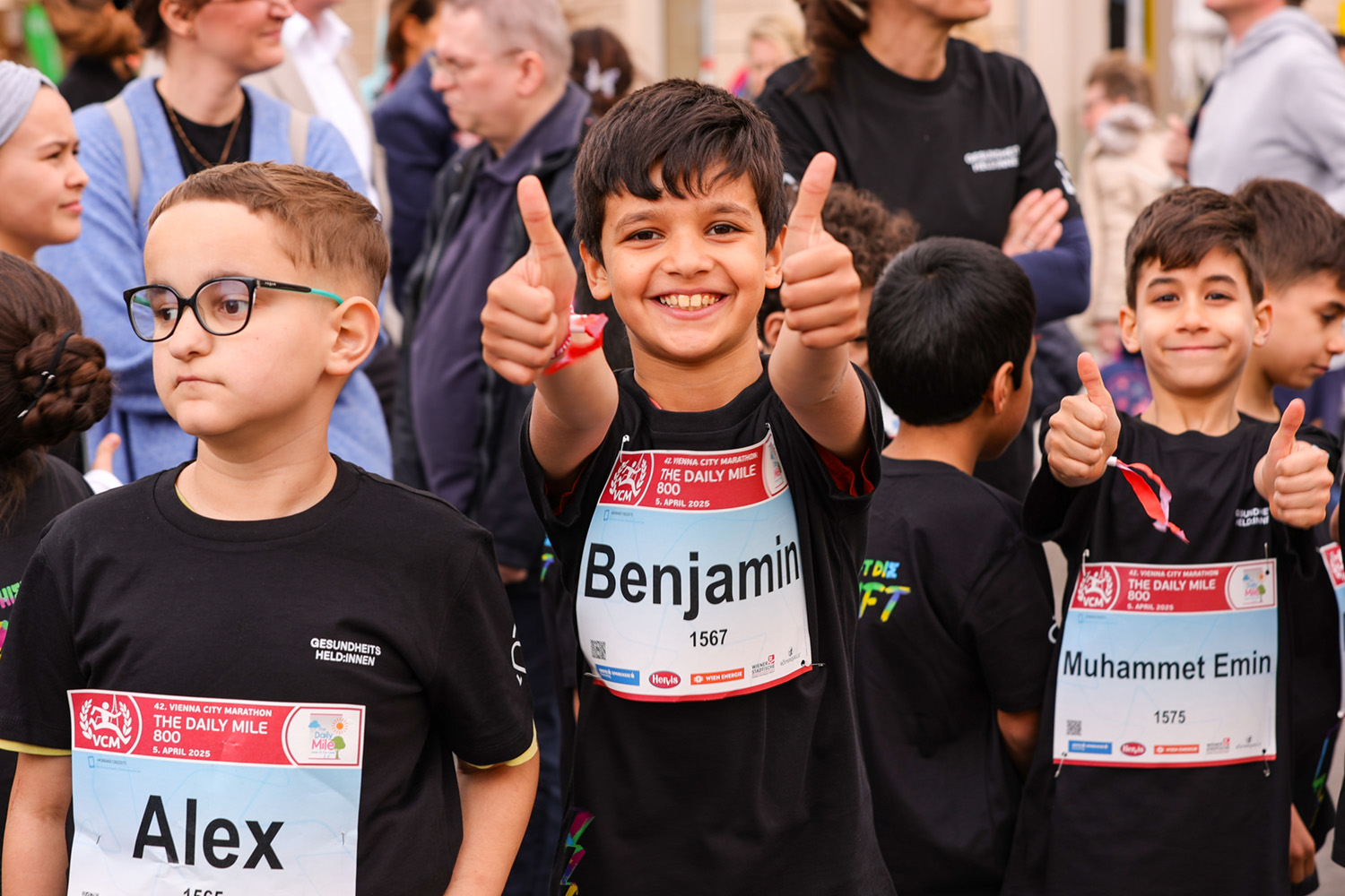 Drei lächelnde Kinder mit Startnummern stehen beim Vienna City Marathon in der Menge. Zwei Jungen strecken beide Daumen hoch.