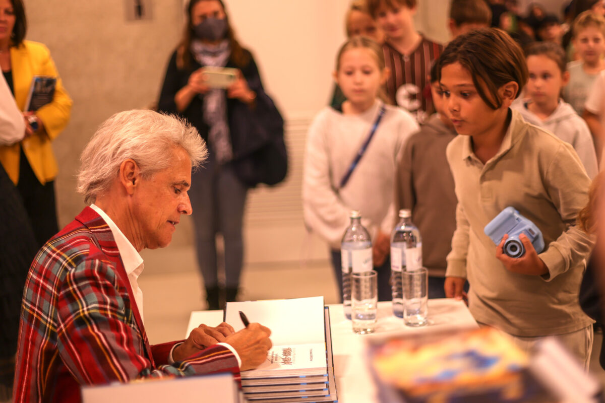 Thomas Brezina signiert ein Buch. Eine Junge sieht ihm begeistert dabei zu.