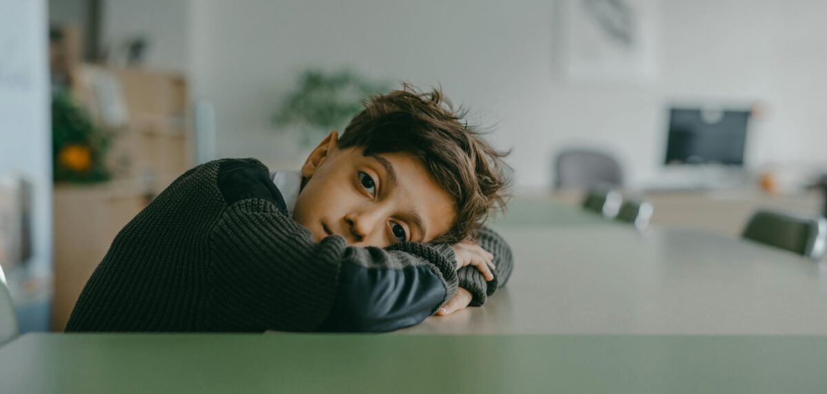 Eine Junge sitzt an einem Tisch. Dabei hat er seinen Kopf auf seinen Armen am Tisch abgelegt und blickt traurig in die Kamera.