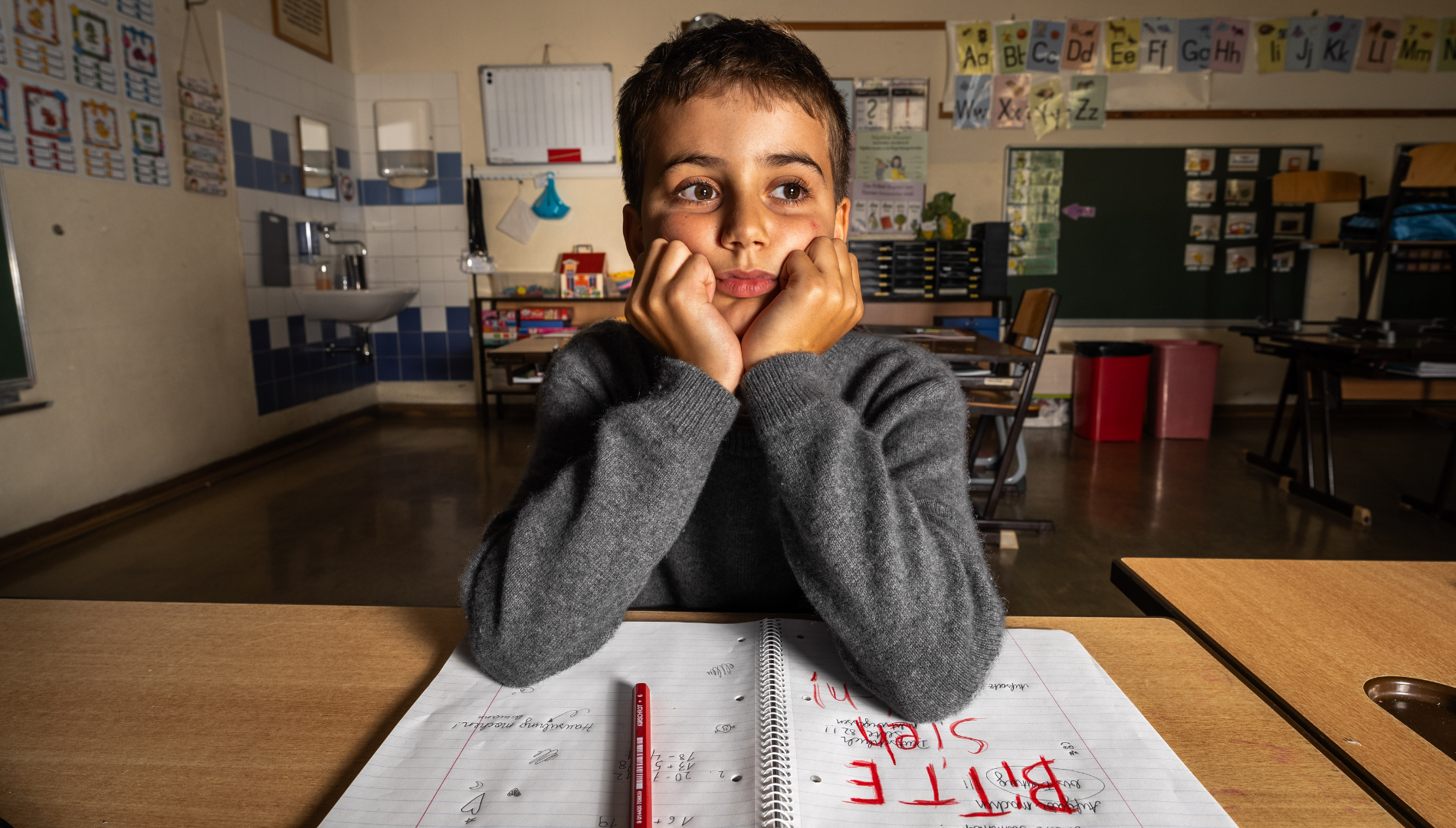Junge sitzt in einem Klassenraum, das Gesicht auf die Hände gestützt, vor einem geöffneten Schulheft mit der Botschaft "Bitte sieh mich".