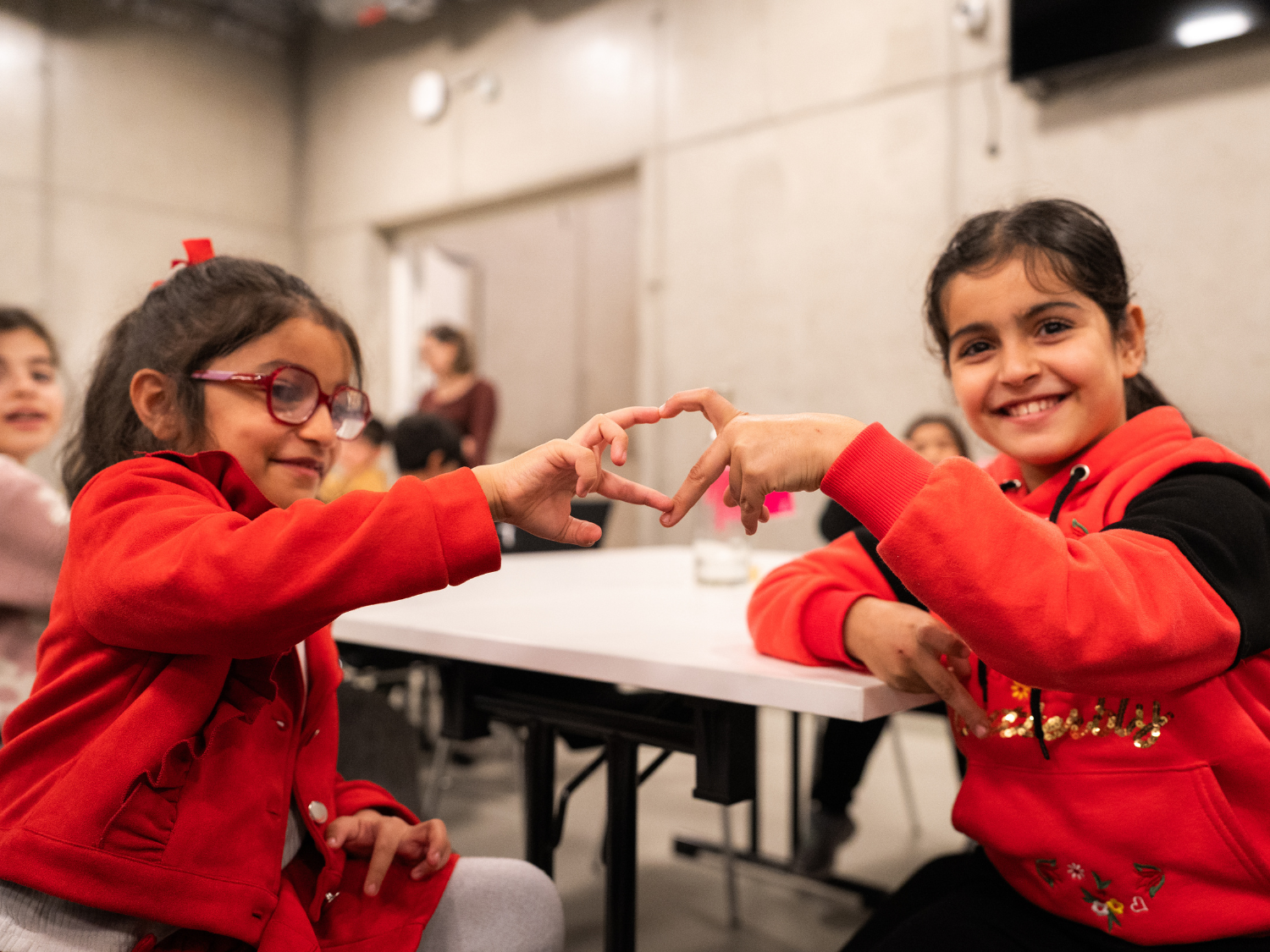 Zwei Mädchen blicken lächelnd in die Kamera und formen mit ihren Händen ein Herz.