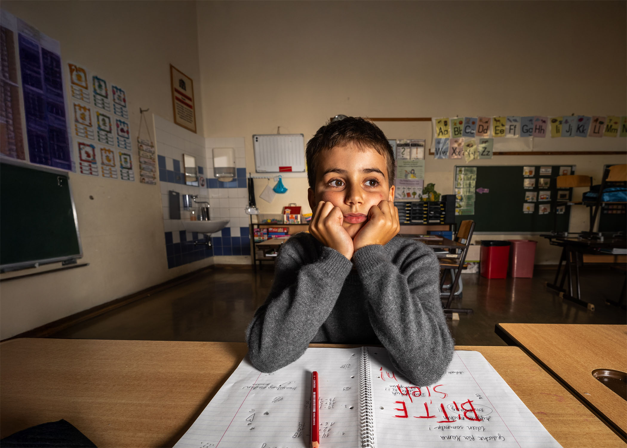 Junge sitzt in einem Klassenraum, das Gesicht auf die Hände gestützt, vor einem geöffneten Schulheft mit der Botschaft "Bitte sieh mich".
