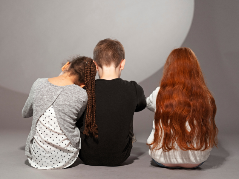 Drei Kinder sitzen mit dem Rücken zu Kamera. Ein Kind lehnt sich an der Schulter des anderen Kind an.