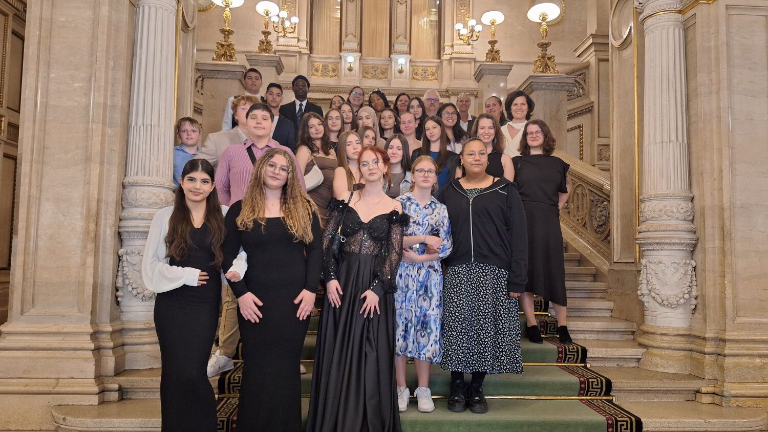 Eine Gruppe junger Menschen in festlicher Kleidung posiert auf einer prunkvollen Treppe in der Wiener Staatsoper.