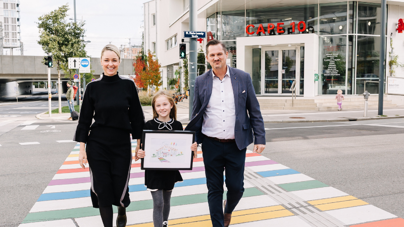 Silvia Bruni, ihre Tochter und Markus Franz gehen mit einer Kinderzeichnung über den Regenbogenzebrastreifen vor CAPE 10.
