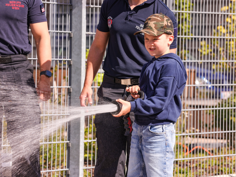 Eine Junge sprüht mit einem Feuerwehrschlauch unter Anweisung der Feuerwehr Wien.